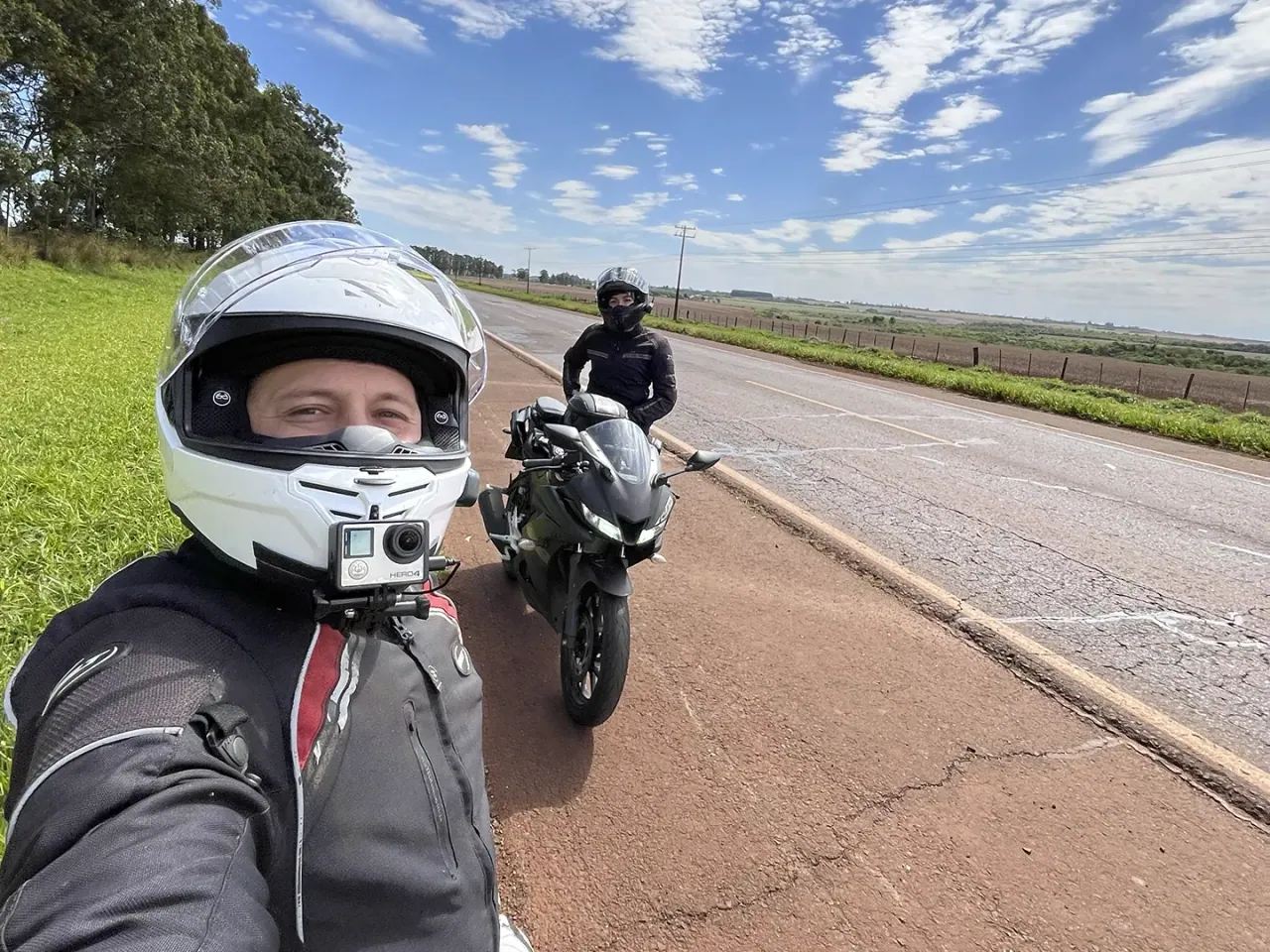 Jeanderson e Ana Paula de capacete durante uma pausa, com a estrada e a paisagem rural ao fundo